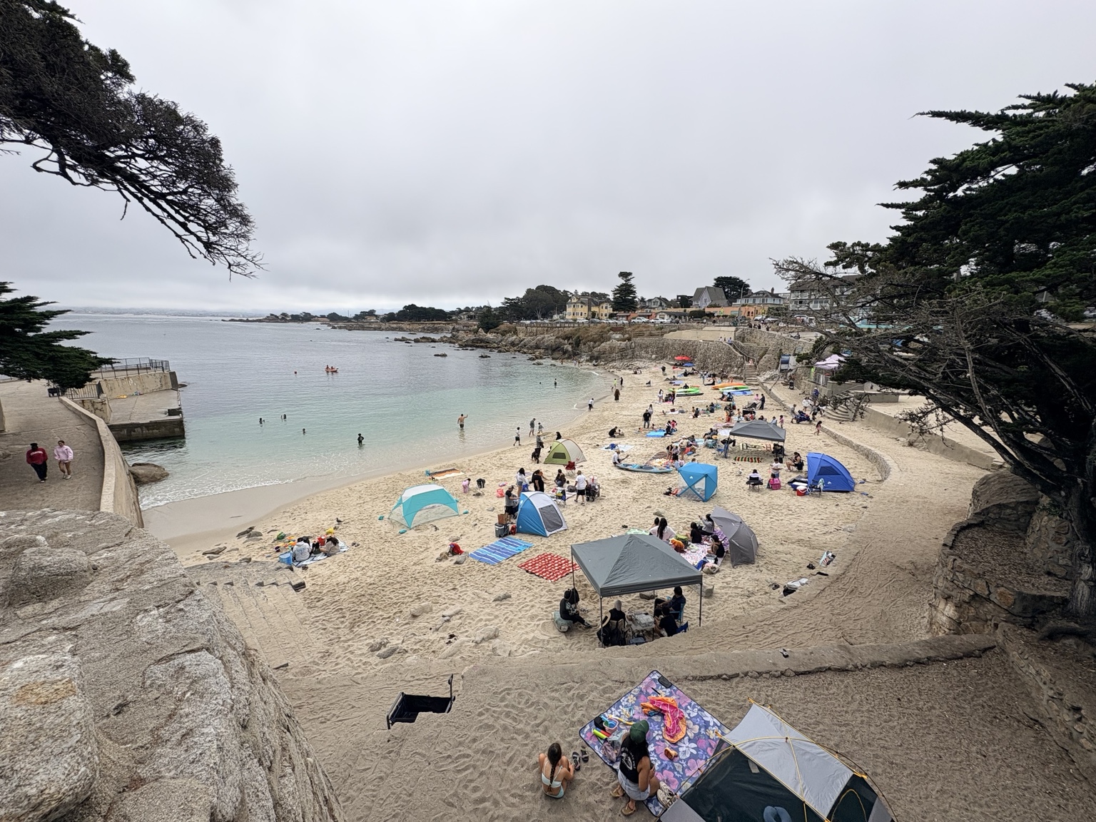 Families, swimmers and beach shelters at Lovers Point Beach beneath an overcast sky on August 8, 2026